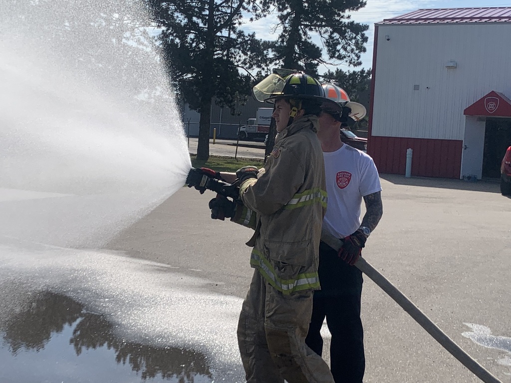 Student in full fire fighter gear holding a hose that is praying water with a fire fighter standing alongside
