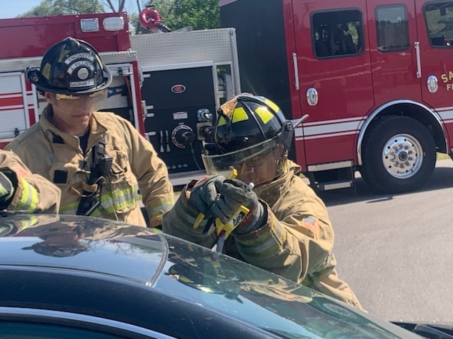 Student fire fighter breaking a car window with an adult fire fighter looking on