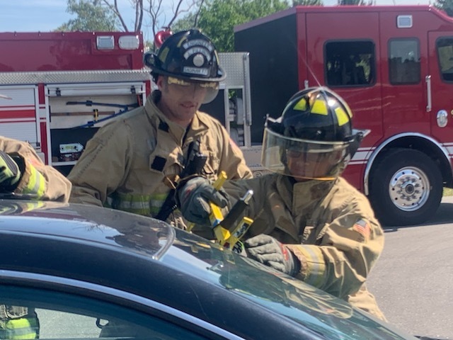 Student fire fighter in gear with an adult fire fighter looking on as they break a car window