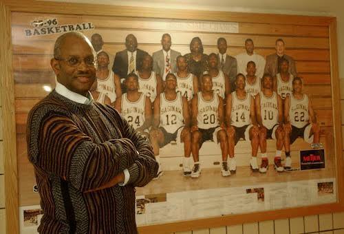Coach T poses in front of a picture of the 1995-96 basketball team