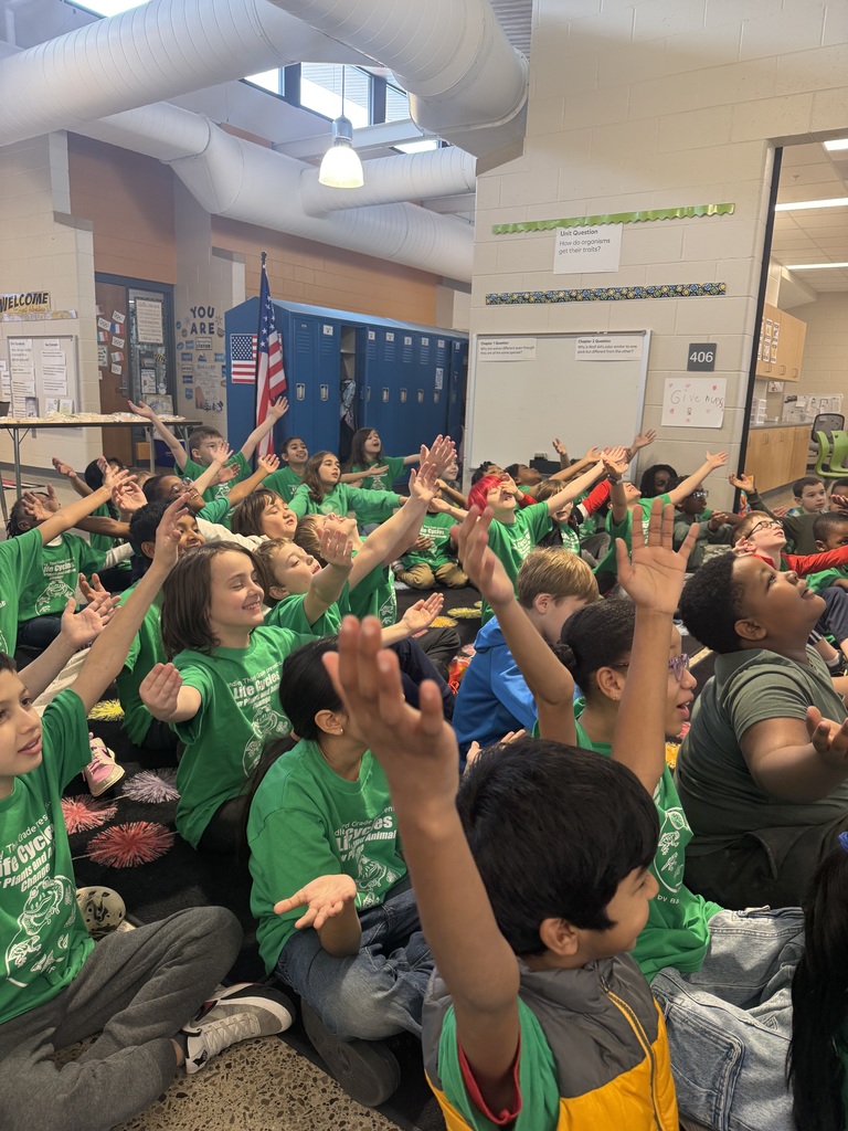 class of students sitting on floor many raising their hands 