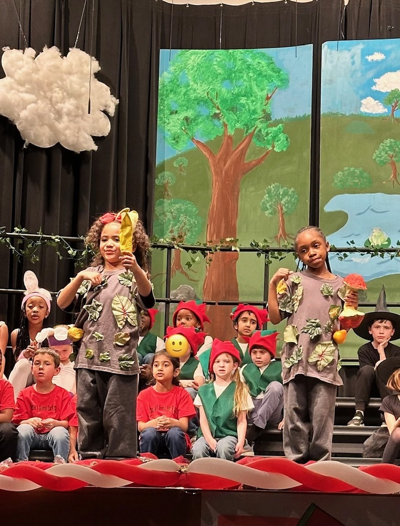 students on stage. 2 standing dressed as trees. brown shirts with leaves