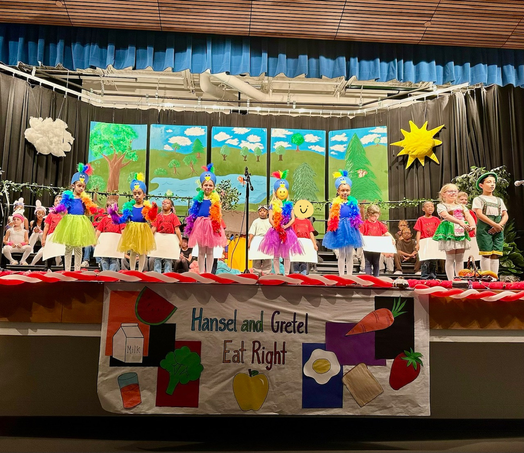 students on stage. students standing are wearing feather boas, colorful shirts, & white skirts