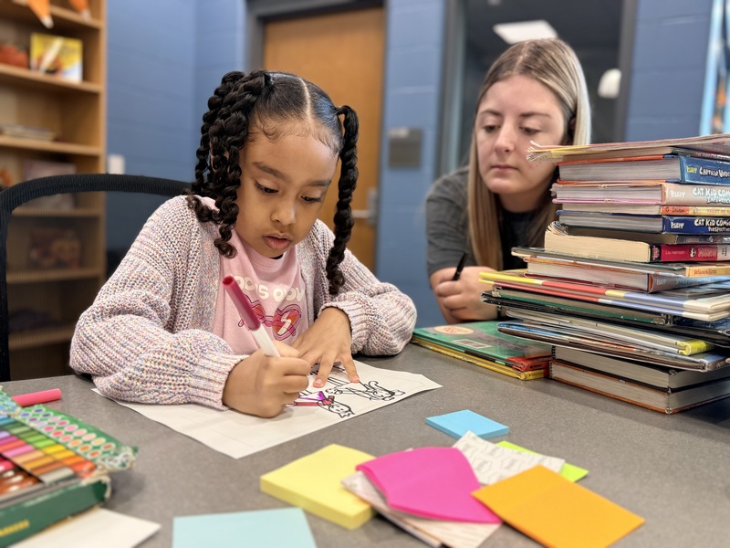 Student writes while sitting next to teacher