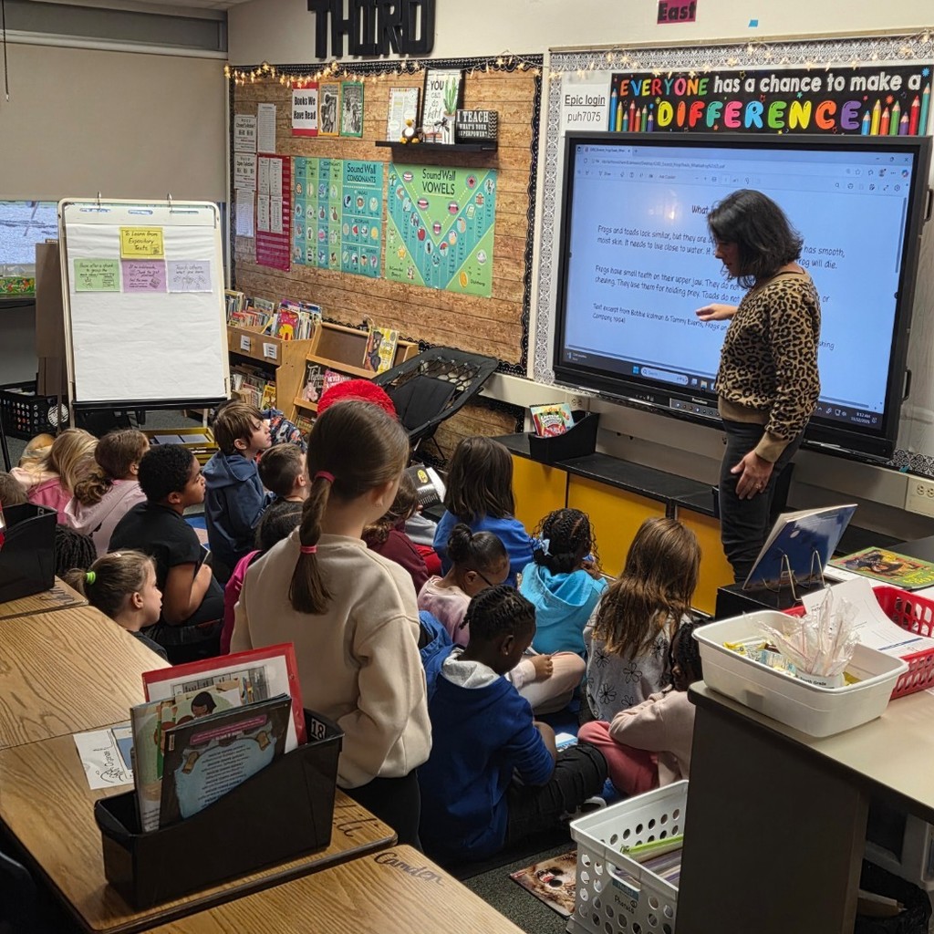 teacher gathered with students at carpet looking at text on big screen