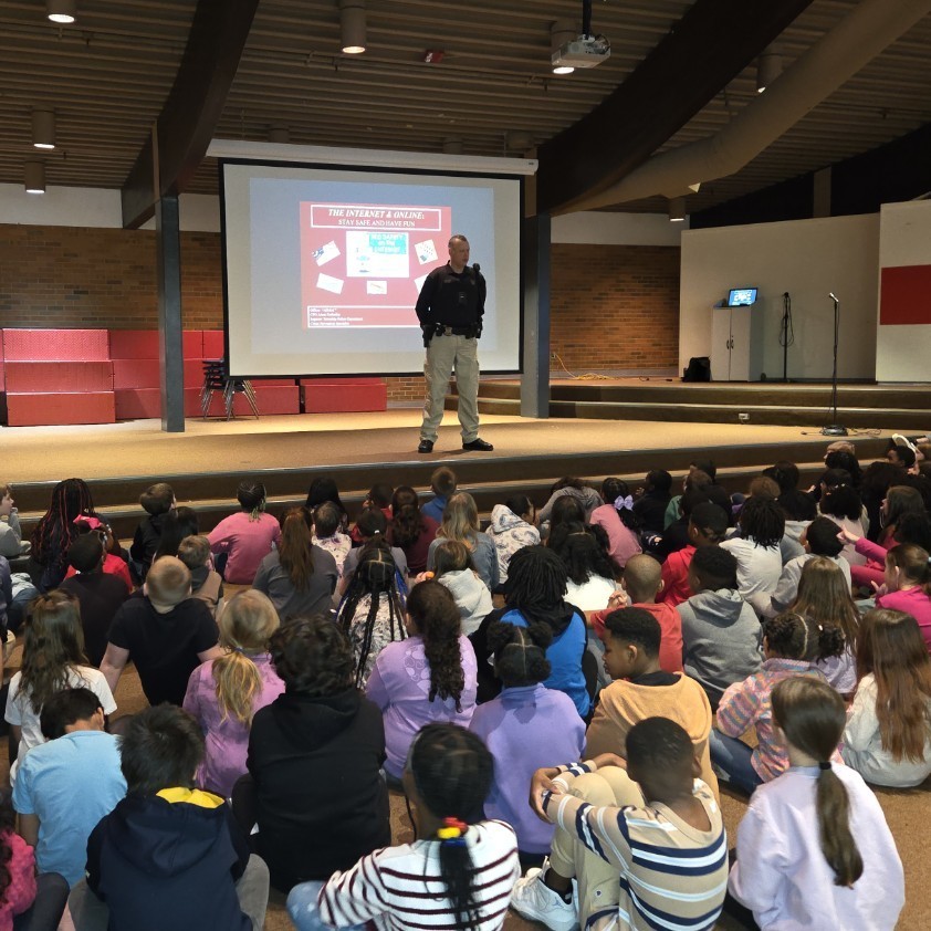 Officer on stage in front of students with displayed slide deck