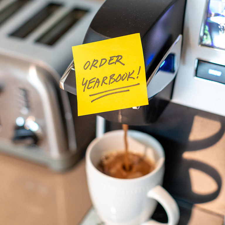 A picture of coffee cup being filled with coffee from a coffee machine. On the machine is a sticky note that says "Order Yearbook!".