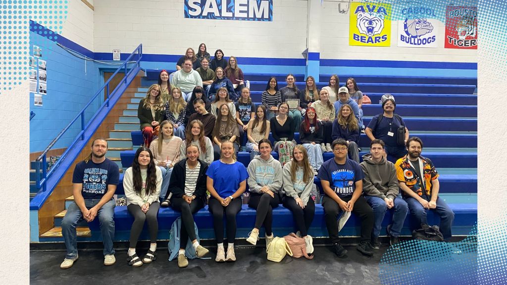 A group of art students and Mr. Nickles and Mr. Bermeister are sitting on bleachers in a gymnasium. The word "SALEM" is written on a banner above them.