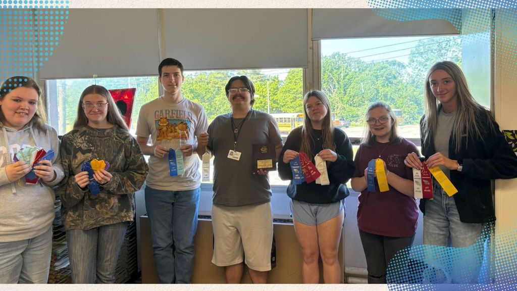 A group of seven people stand in front of a window, holding awards and ribbons.