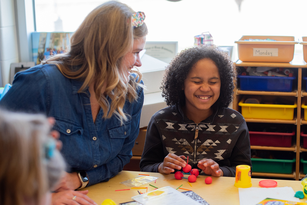 Girl playing with plato and a teacher sitting next to her.