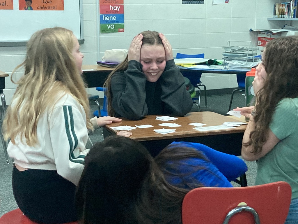 Four students sit around a classroom desk during a fun activity; one student in the center has her hands on her head with a playful, frustrated expression while looking at game cards.