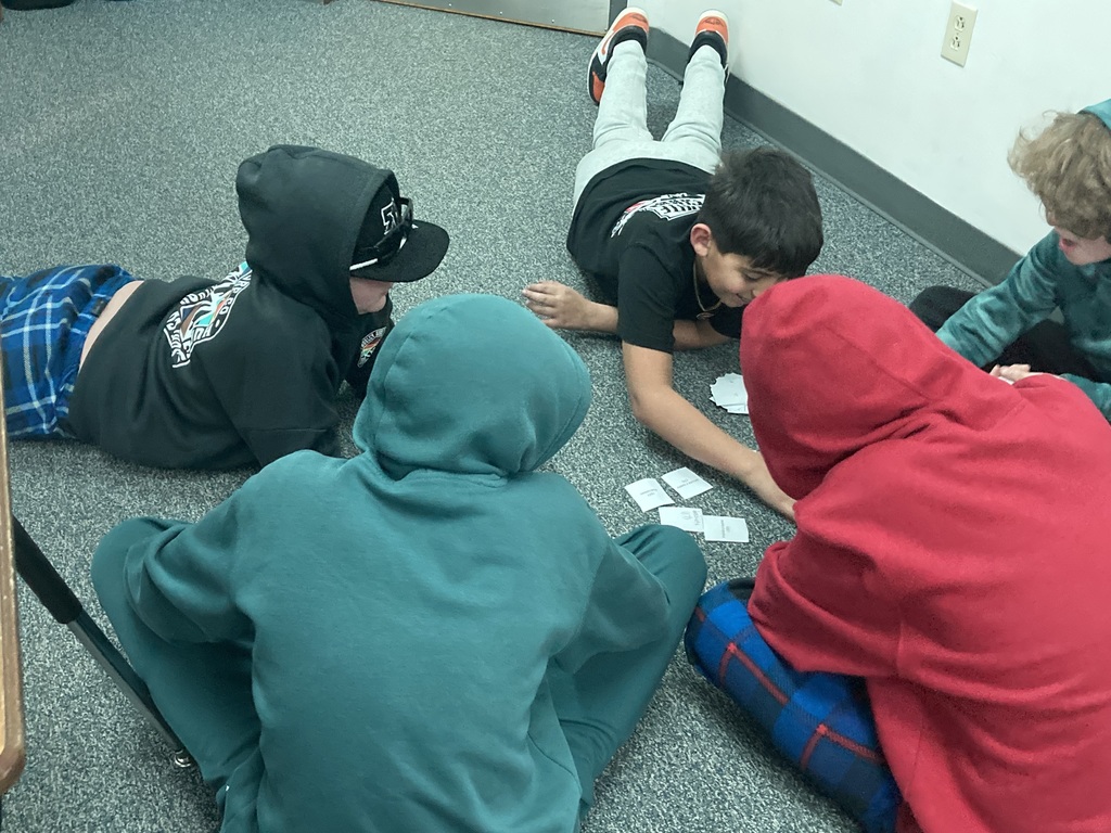 A group of students in hooded sweatshirts and casual wear collaborate on the floor of a classroom, focused on arranging small vocabulary slips during a group activity in Spanish.