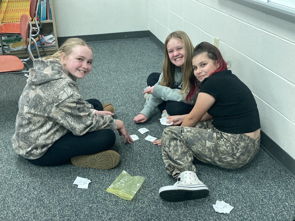 Three middle school girls sit in a circle on a grey carpeted classroom floor, smiling and laughing while playing a learning game with small white cards.