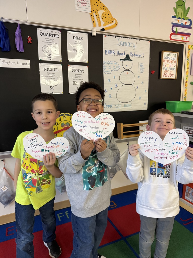 Three boys grin while displaying paper hearts during a classroom activity. The hearts are covered in colorful writing with words like "respectful," "joker," and "cool." One student wears a bright yellow Pokémon shirt, and they are positioned in front of a bulletin board displaying lessons on US currency.