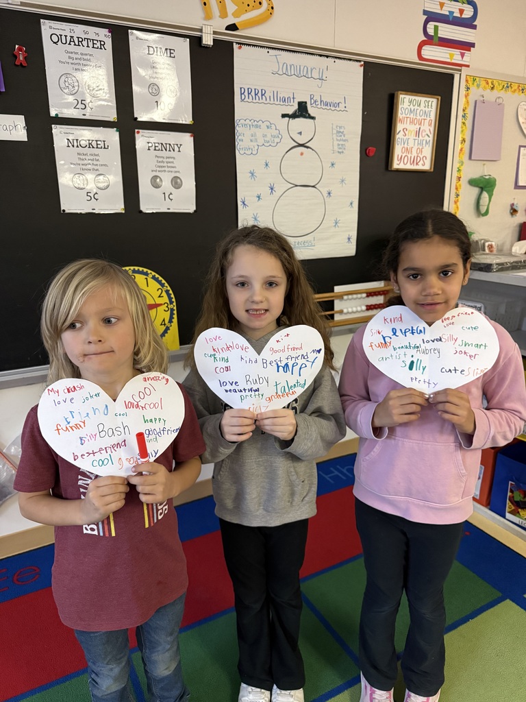 Three young kids smile for a photo while holding decorated paper hearts in their classroom. The hearts feature names like Aubrey, Ruby, and Bash surrounded by kind words such as "best friend," "talented," and "helpful." The classroom background shows a January-themed "Brilliant Behavior" chart.