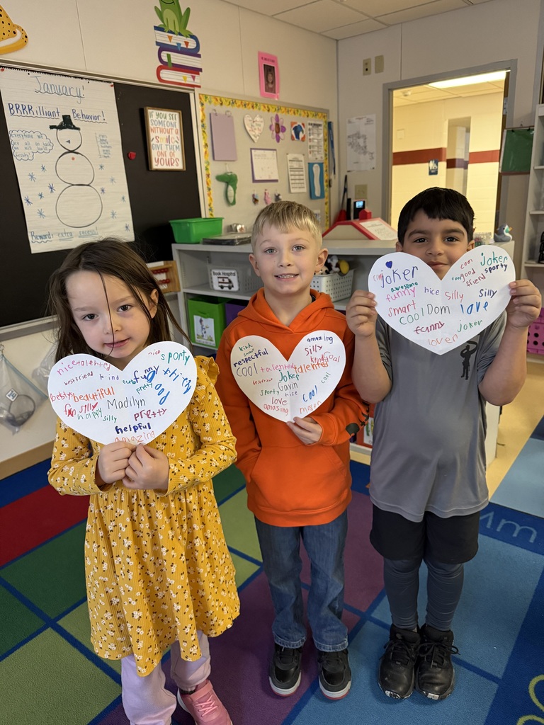 Three elementary students stand in a row, each holding a large white paper heart with their names—Dom, Gavin, and Madilyn—written in the center. The hearts are decorated with colorful handwritten words describing their positive traits. They are standing on a vibrant, lettered classroom rug.