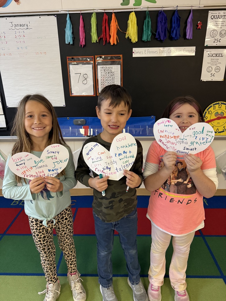 Three students pose in a classroom holding paper hearts that highlight positive character traits. The hearts include names like Sophia, Teddy, and Joanna, surrounded by words like "grateful," "responsible," and "amazing." The classroom wall behind them features a "January" math and behavior chart.