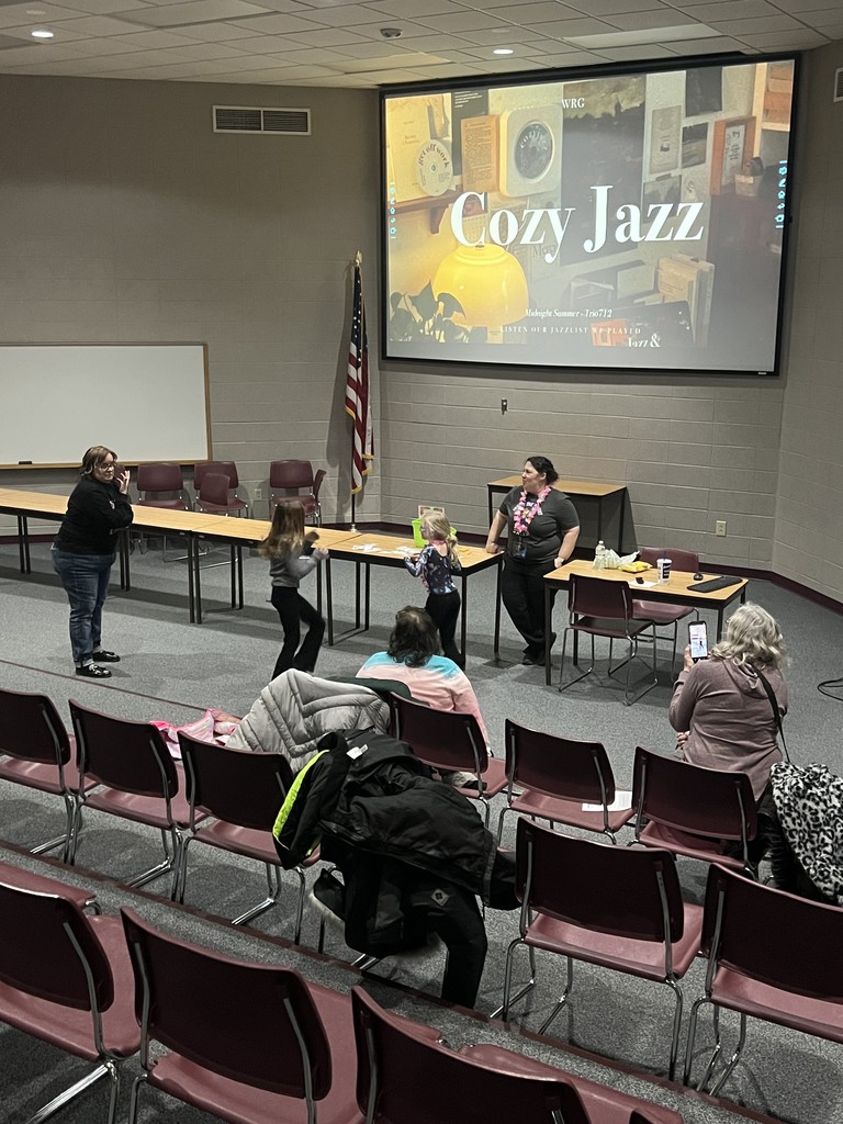 A school presentation room with a large screen displaying "Cozy Jazz" while students and staff playing charades near rows of chairs.