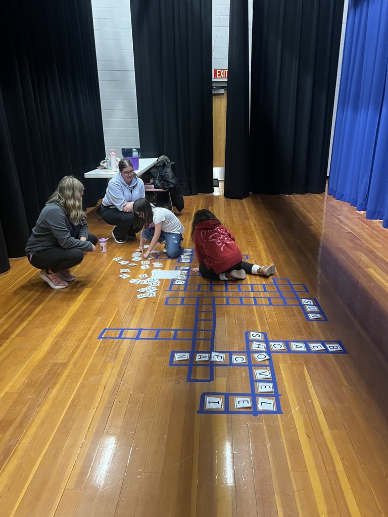 Families sitting on a wooden stage floor playing a giant crossword puzzle game made with blue tape and letter cards.