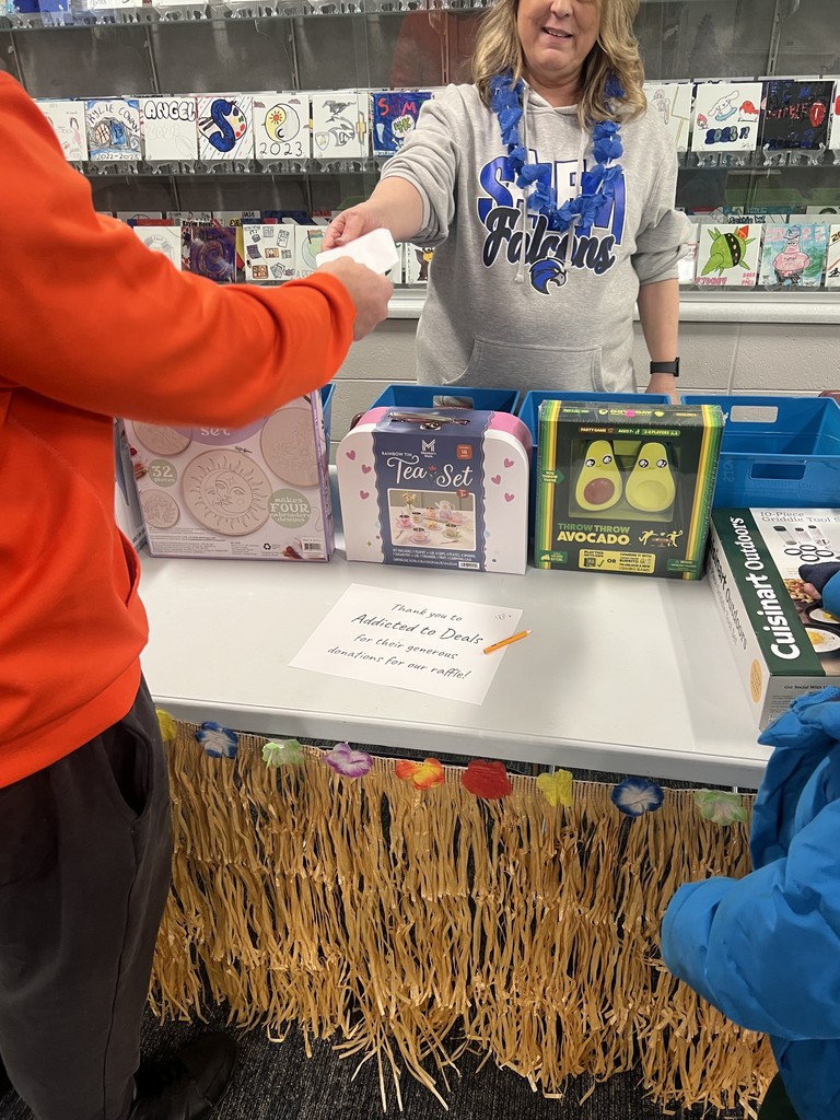 A volunteer at a raffle table decorated with a grass skirt and flowers, showing prizes like a "Throw Throw Avocado" game and tea set.