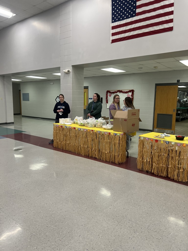 Volunteers setting up a snack station at a table decorated with a tropical grass skirt in a school hallway near an American flag.