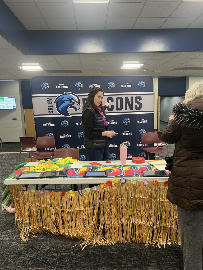 A staff member wearing a lei stands behind a table decorated with a grass skirt in front of a Salem Falcons school backdrop.