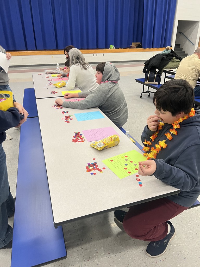 A student wearing a floral lei plays a bingo game at a long cafeteria table with colorful bingo cards and plastic markers.