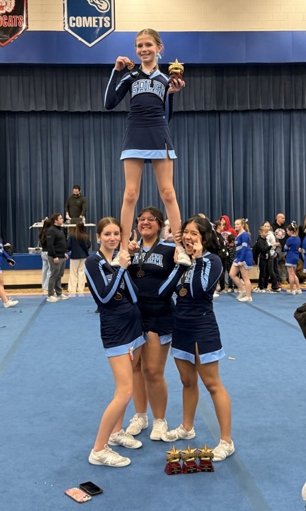 Four Salem cheerleaders performing a stunt in a gymnasium, with the flyer holding a trophy and medal after a competition.