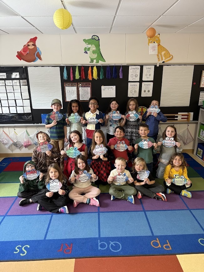 A group of elementary students sitting and standing in a classroom, holding up colorful, hand-drawn monster crafts in front of a chalkboard.