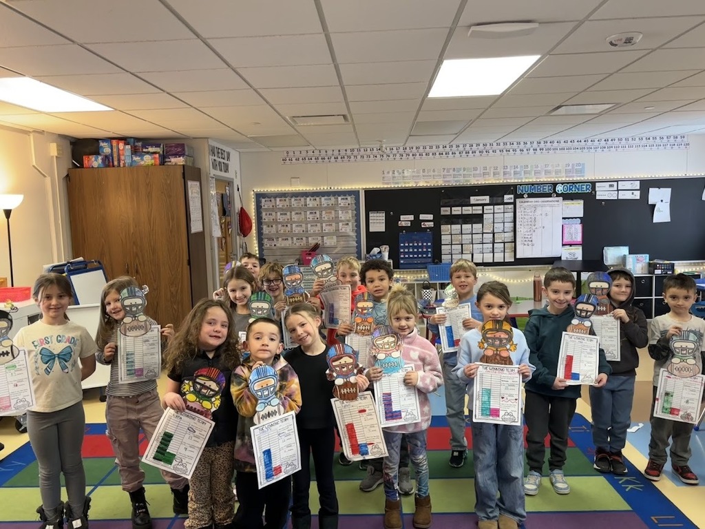 A classroom of students standing on a colorful rug, proudly holding up their "The Winning Team" football-themed math and coloring projects.