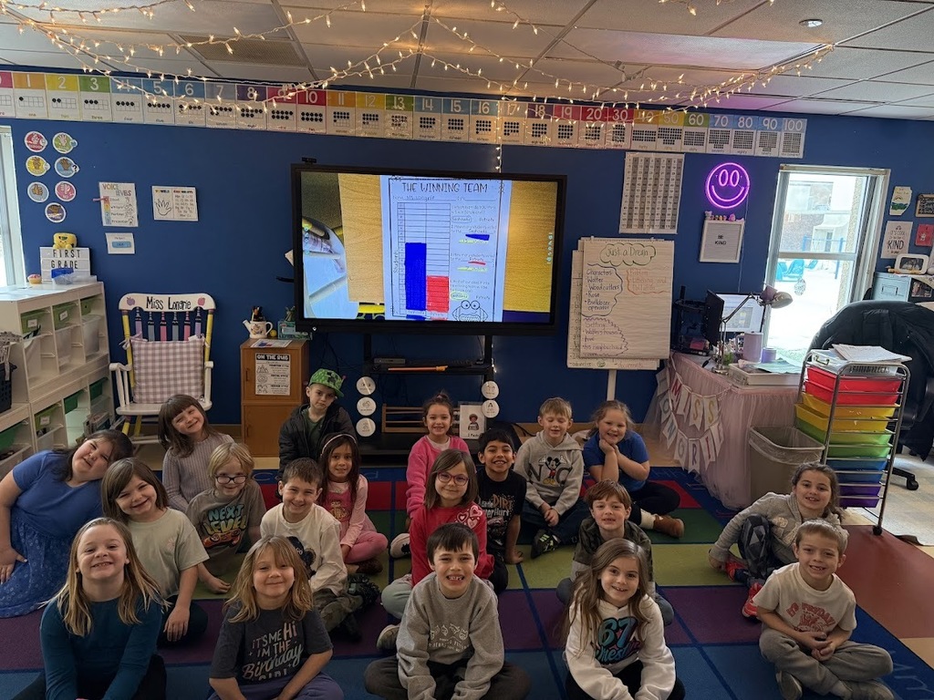 A group of primary students sitting on a classroom rug in front of a large screen displaying a "The Winning Team" bar graph lesson.