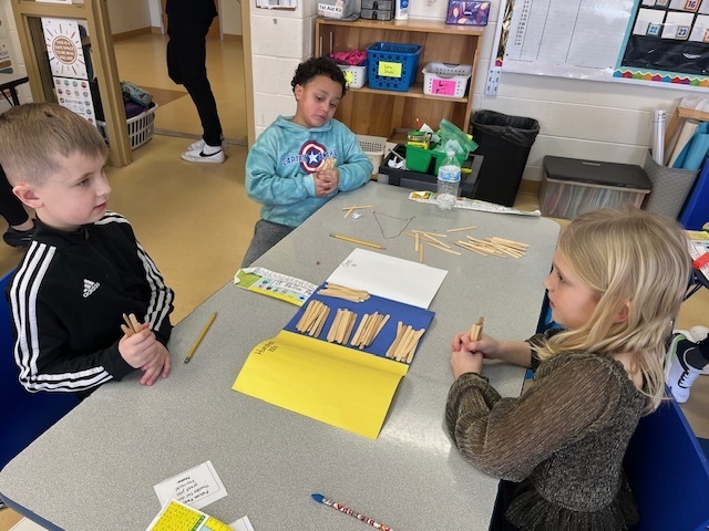 Three students collaborate at a table, using bundles of wooden sticks on a place-value mat to represent different numbers during a math lesson.