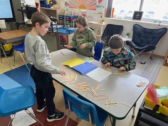 Three students collaborate at a table, using bundles of wooden sticks on a place-value mat to represent different numbers during a math lesson.