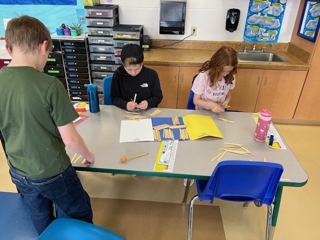 Two students sit at a table sorting bundles of craft sticks onto blue and yellow mats labeled for place value while a third student stands nearby.