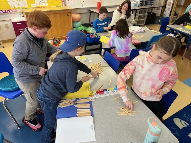 Three young students stand around a classroom table, sorting wooden craft sticks into piles on colored paper to practice counting and place value.