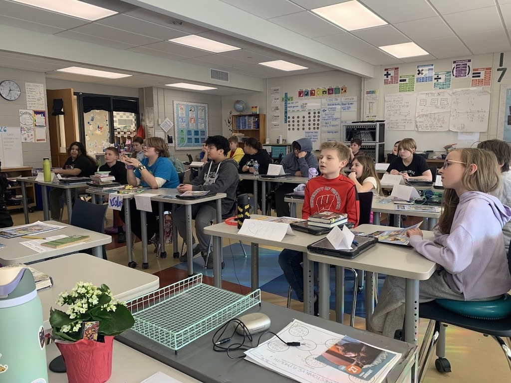 A classroom of elementary students sitting at desks, listening attentively to a lesson. A plant and classroom supplies are in the foreground.