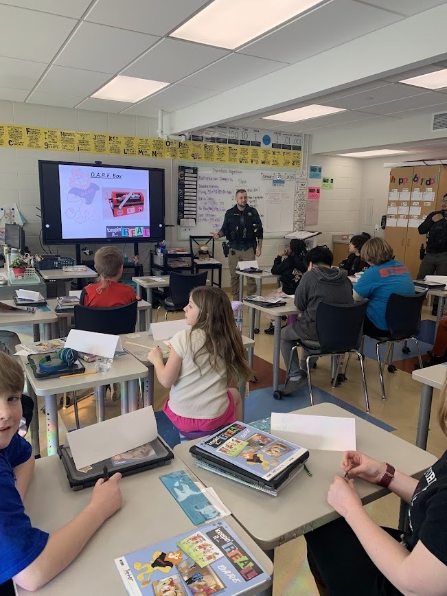 A police officer gives a D.A.R.E. presentation to a class of students. A digital screen displays the "keepin' it REAL" logo.