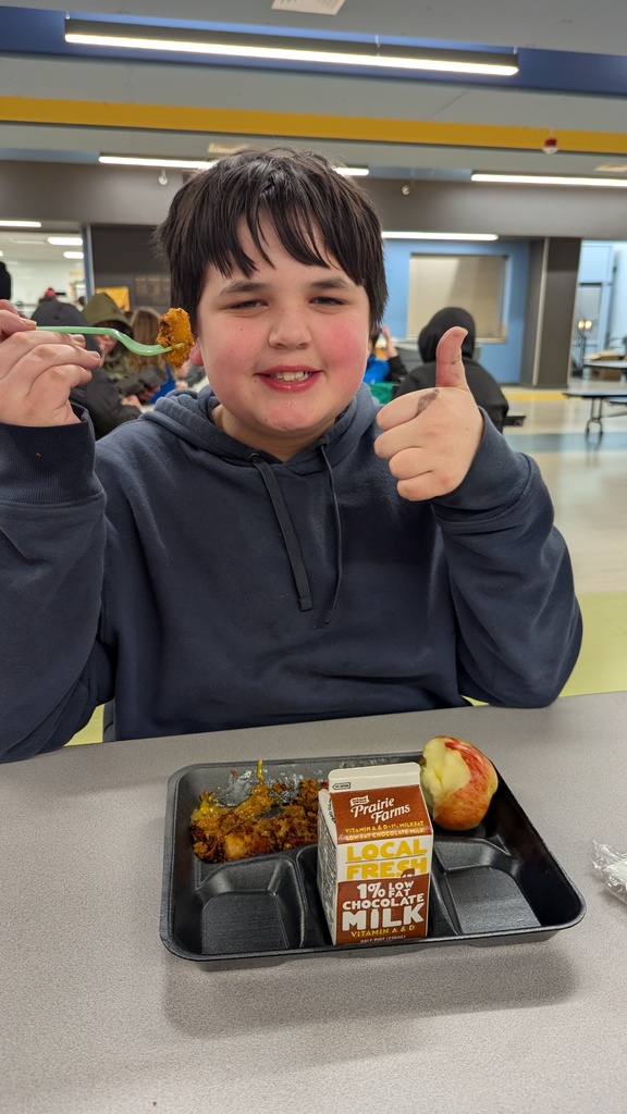 A student gives a thumbs up while enjoying a school lunch of loaded tater tots, an apple, and chocolate milk.