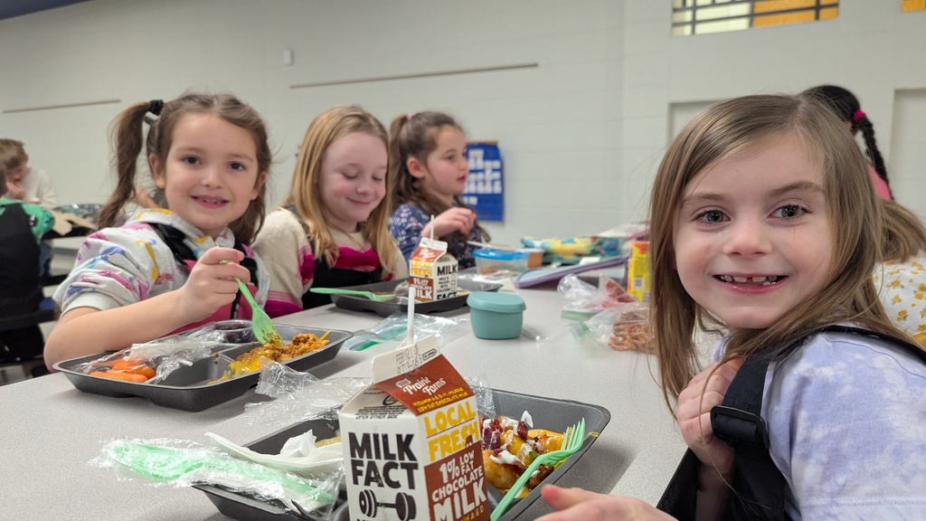 A group of elementary students sitting together and smiling while eating lunch in a bright school cafeteria.
