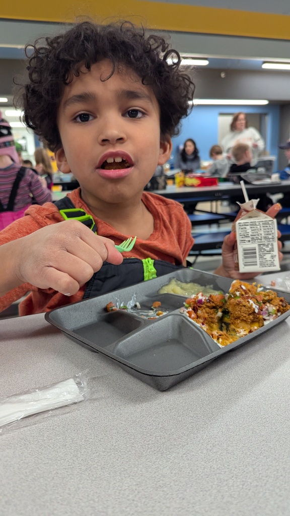 A young student with curly hair eating a school lunch of loaded tater tots and applesauce while holding a small milk carton.