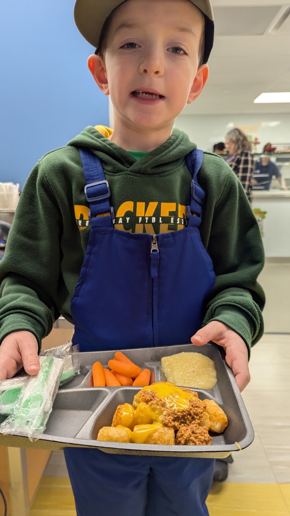 A young student in a Green Bay Packers sweatshirt holding a school lunch tray with tater tot nachos, baby carrots, and applesauce