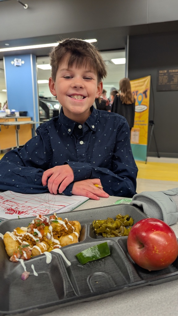 A smiling student in a blue button-down shirt sits with a school lunch tray of loaded tots, jalapeños, and a red apple.