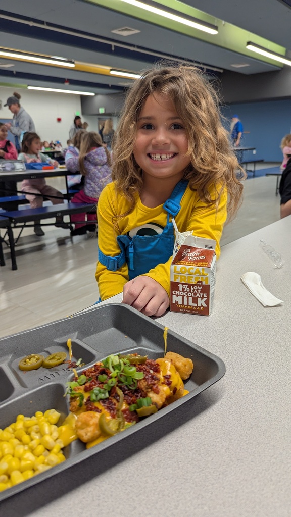 A smiling student with curly hair sitting at a cafeteria table with a carton of chocolate milk and a tray of loaded tater tots and corn.