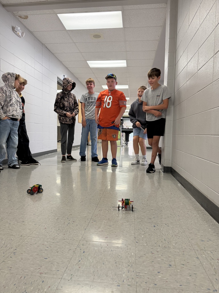 A group of middle school students are in the hallway watching 2 cars race that were created in PLTW Robotics class.