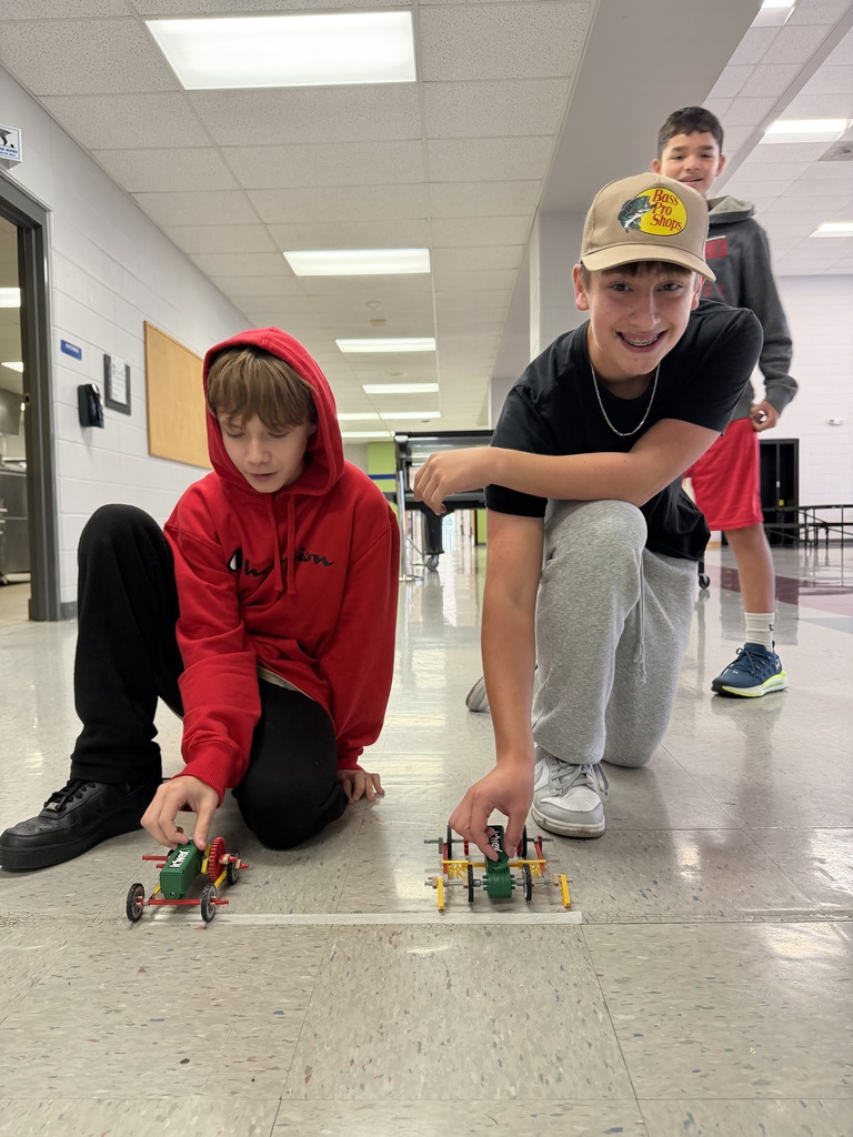 2 students are racing cars that they created in PLTW robotics in the school hallway.  
