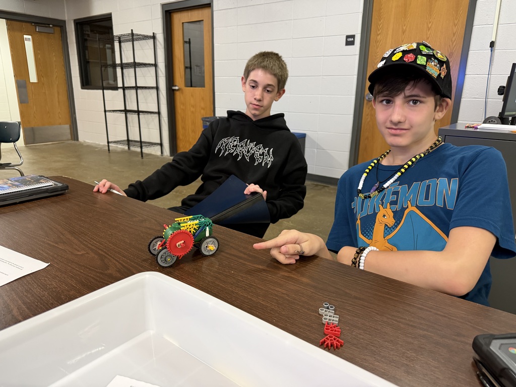 2 students sit at a table and show off the car that they made during PLTW Robitics class