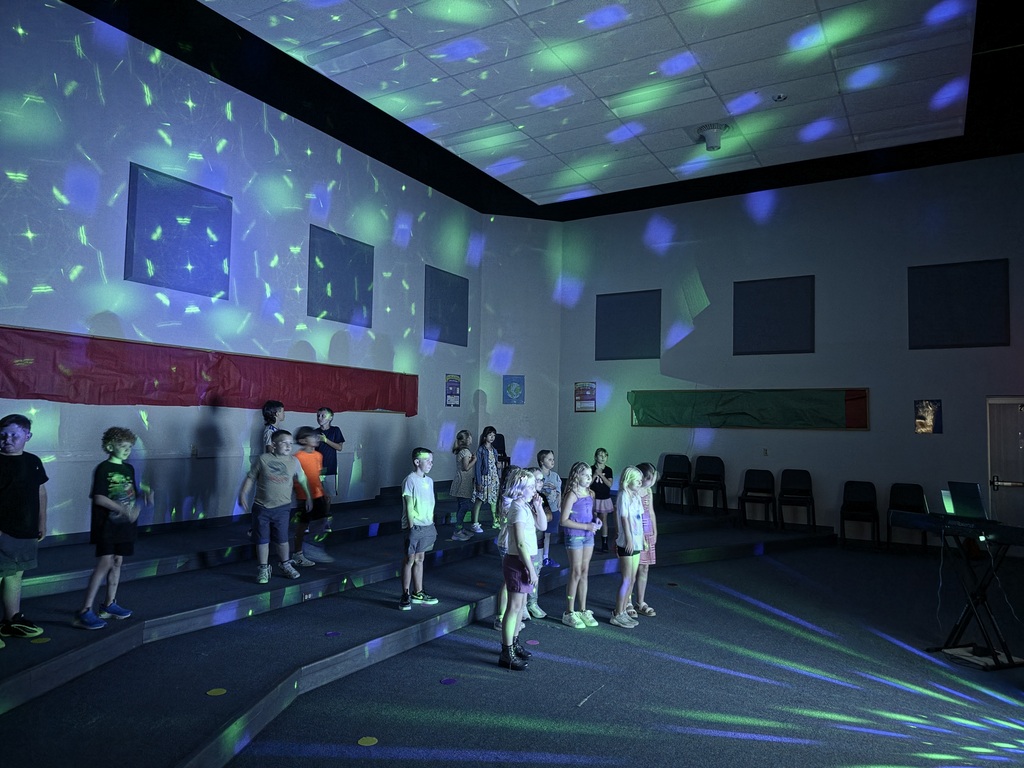 A group of elementary students stands on tiered risers in a dimly lit music room, practicing singing under bright blue, green, and white patterned stage lighting.