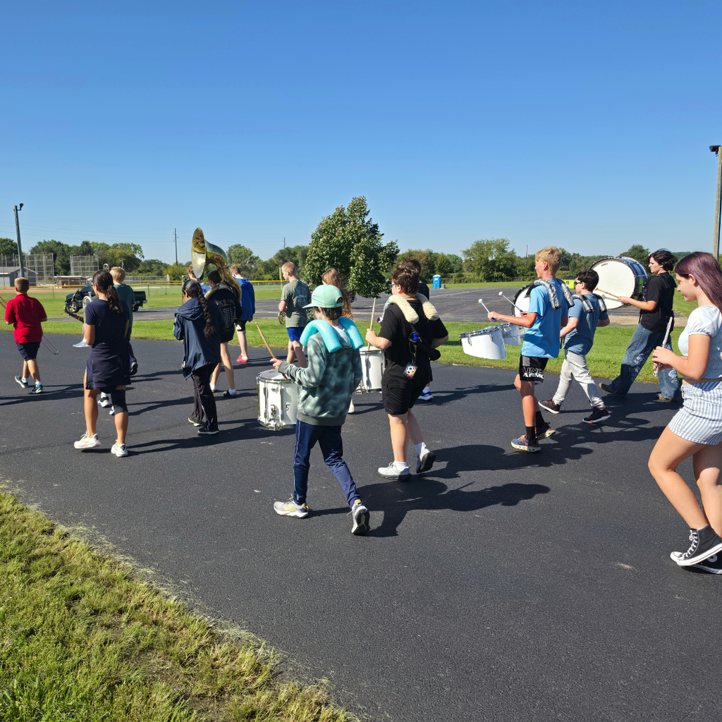 A group of middle school band students marches down a paved outdoor path on a sunny day with clear blue skies, carrying and playing percussion, brass, and woodwind instruments during practice.