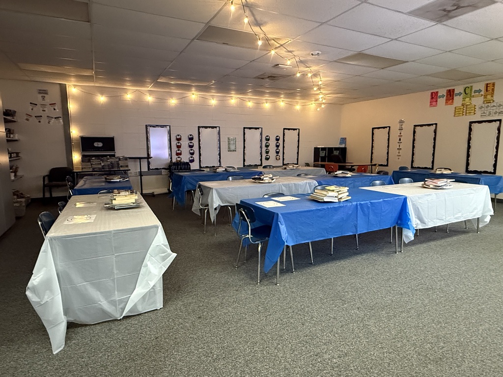 An empty classroom decorated with string lights, set up for a book tasting with tables covered in blue and white tablecloths and stacked books.