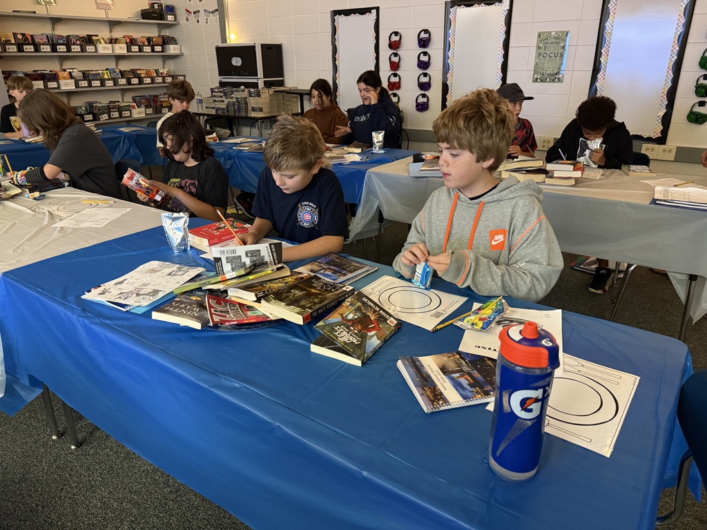Classroom scene showing students seated at long tables decorated with blue covers, browsing books and completing reading activities.